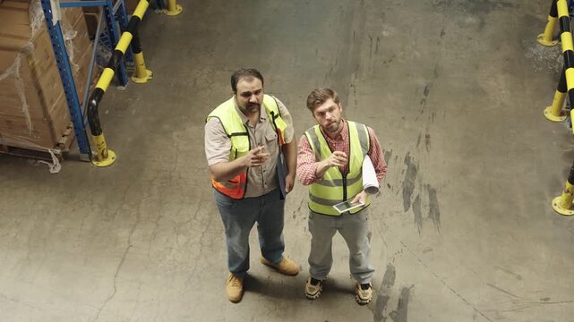 Two Caucasian men collaborate during warehouse inspection, discussing logistics amidst stacked boxes and safety barriers within darkened industrial setting for quality control assessment.