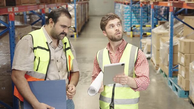 Two men collaborate reviewing warehouse inventory using tablet and documents within brightly lit industrial facility filled with stacked boxes; supply chain management solutions for logistics optimiza