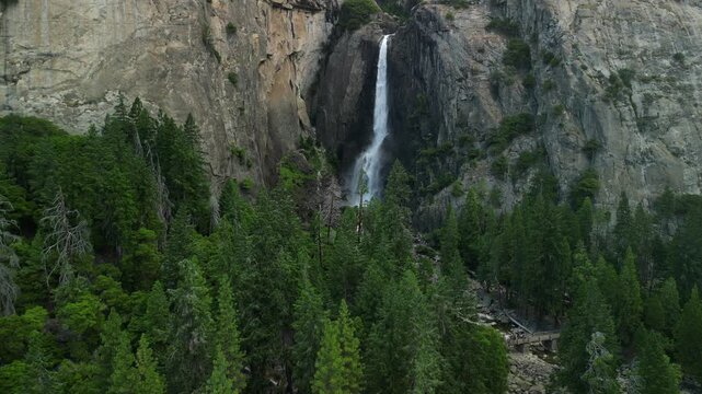 Aerial shot of Yosemite Valley featuring cascading waterfall surrounded by granite cliffs, dense forest, and dramatic mountain landscape in California&rsquo;s Sierra Nevada.