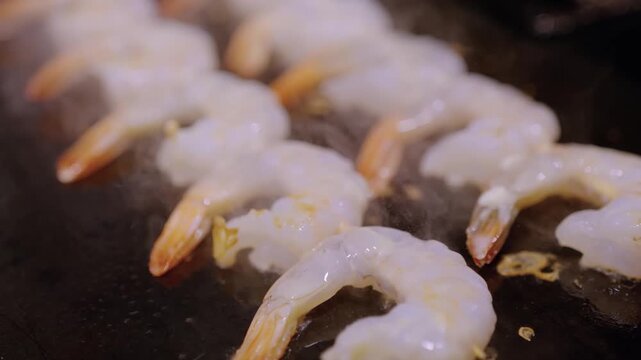 Close-up shot of peeled and deveined raw shrimp being cooked on a hot griddle or Blackstone. The footage shows the translucent flesh starting to turn opaque and pink as they are seared in hot oil.