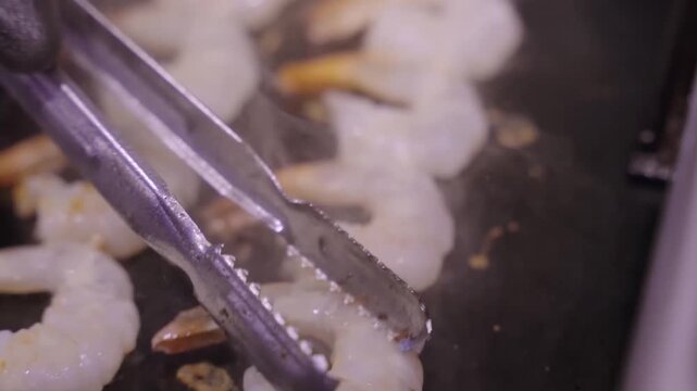 A high-quality close-up shot showing rows of fresh, peeled prawns being cooked on a professional black flat-top grill. Steam rises as the seafood sizzles, and a pair of metal tongs is used to carefull