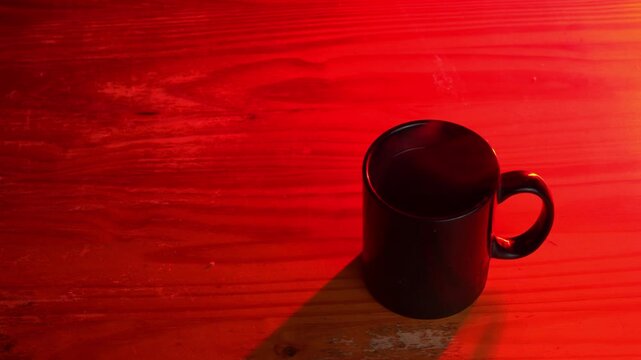 Top angle close up of steaming black mug on a red lit wooden table, Hot beverage with wisps of steam on a rustic wood surface in red neon light