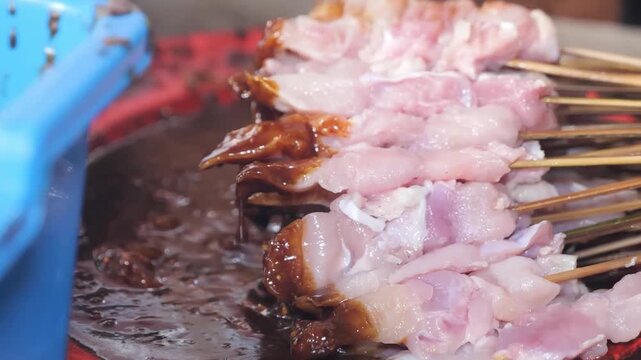 A detailed close-up shot of raw marinated pork skewers being dipped or held over a thick, dark spicy sauce at a street food stall. The footage captures the preparation of traditional Asian BBQ, often 