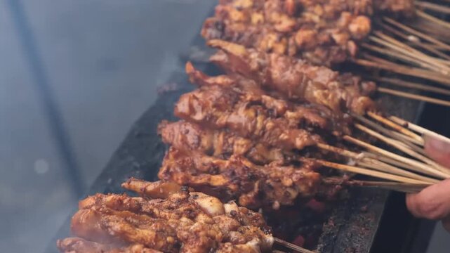 Close-up of traditional Indonesian chicken satay skewers being grilled over glowing charcoal embers. The marinated meat is seasoned with authentic spices and cooked on a long outdoor grill, producing 