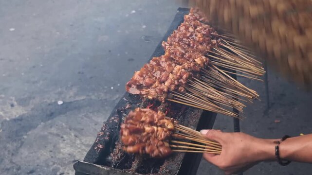 A close-up of a street food vendor grilling rows of seasoned chicken satay (Sate Ayam) on bamboo skewers over a traditional charcoal grill. Thick smoke rises from the glowing embers as the meat carame