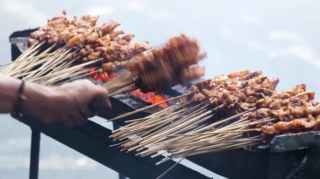 A close-up shot of numerous chicken satay skewers (Sate Ayam) being grilled by a street food vendor over hot charcoal. The footage captures the traditional cooking process with rising smoke and the ve