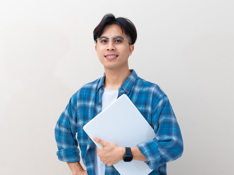 A smiling young Asian IT professional or freelancer standing while holding a closed laptop against a plain background.