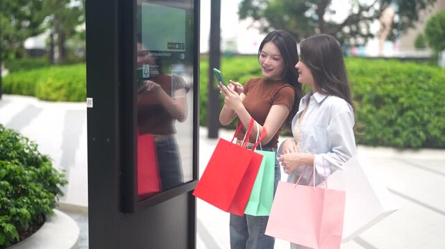 Two happy asian female friends holding shopping bags scanning QR codes on digital city kiosk with smartphone, Smart urban lifestyle, modern technology, mobile app navigation, new consumerism concepts
