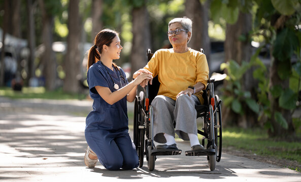 Caring Asian Nurse Kneeling and Holding Hands with Senior Woman in Wheelchair
