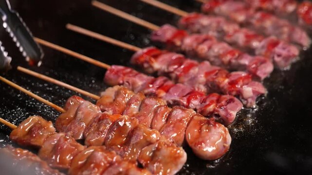 close-up shot of seasoned chicken meat skewers (Yakitori) sizzling on a traditional Japanese charcoal grill. Steam and smoke rise from the hot grill as metal tongs adjust the skewers. Authentic Japane
