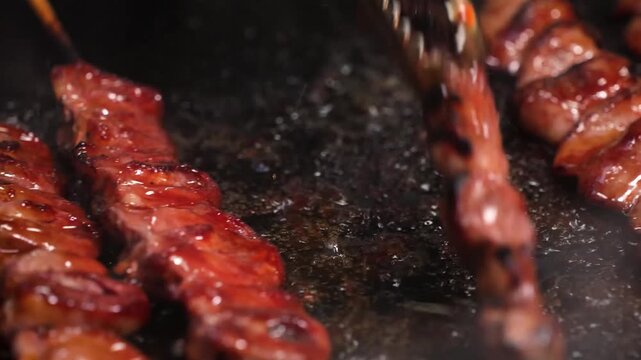 close-up shot of marinated pork belly pieces on bamboo skewers being grilled over charcoal. The meat is glazed with a sweet and savory Filipino-style barbecue sauce, showing a glistening, caramelised 