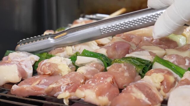 Footage of a chef preparing traditional Japanese yakitori (Negima) by seasoning raw chicken thigh and scallion skewers with salt over a professional grill in a kitchen setting.