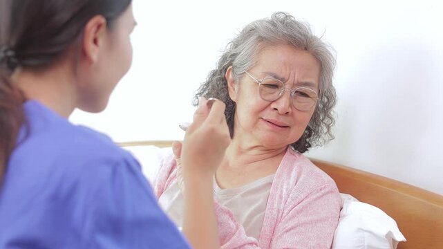 Senior Asian woman refusing food from a caregiver in bed. Elderly patient showing loss of appetite, looking unhappy and turning away from the spoon. Professional home healthcare concept.