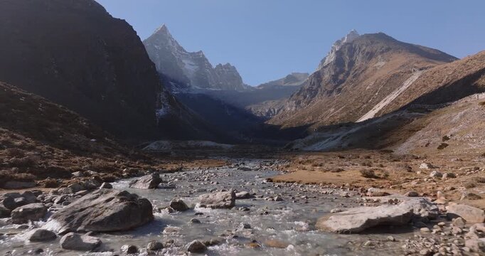 Everest region, Nepal, aerial push in shot toward a flowing freshwater stream with a rocky bottom winding through an alpine valley beneath Mount Cholatse and Taboche peaks.