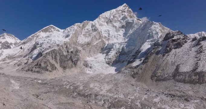 Gorakshep Hamlet, Everest Nepal, drone shot of Himalayan crows flying across a rugged glacial moraine beneath the towering snow capped peak of Nuptse in the Everest region.
