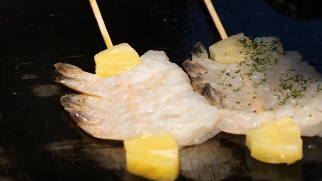 Fresh shrimp skewers with pineapple chunks sizzle on a hot flat-top griddle, showcasing a vibrant street food or kitchen atmosphere. The peeled raw shrimp are neatly arranged on bamboo sticks with bri