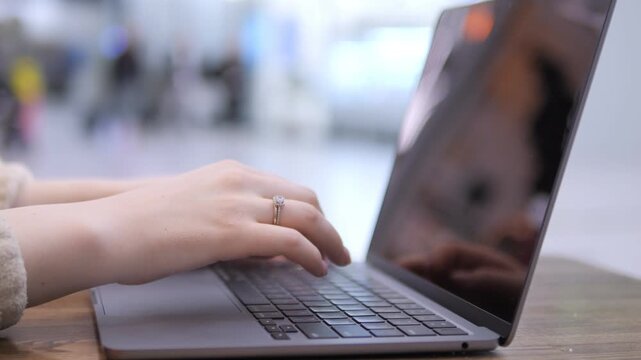 Hands type on laptop in public space. Laptop screen reflects surroundings. Background is blurred with people passing by