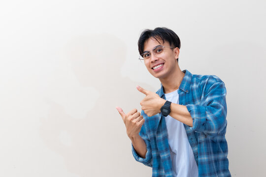 Smiling young Asian man wearing glasses and a plaid shirt pointing his thumbs sideways at copy space against a plain background
