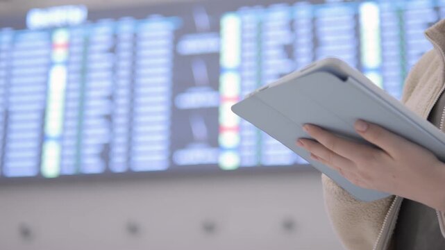 Person holds tablet near airport departure board. Background shows flight information screens. Hands visible, wearing beige coat. No animals or plants shown