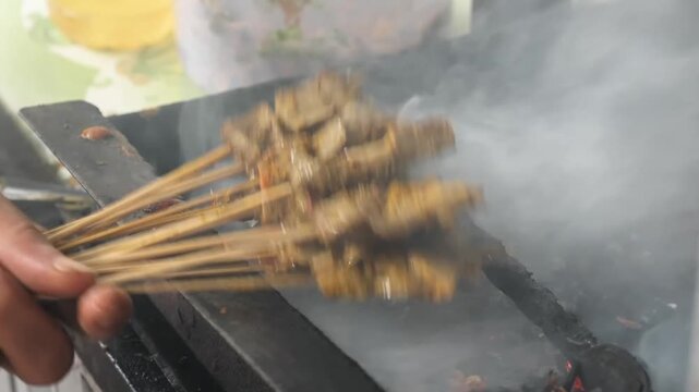 Close-up footage of many satay (sate) skewers being grilled on a traditional charcoal pit. The scene captures seasoned meat skewers sizzling over hot coals with flavorful white smoke rising, represent
