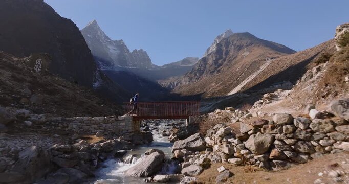 Everest region, Nepal, aerial push-in shot of a trekker with a backpack crossing a red suspension bridge over a rocky mountain river beneath Mount Cholatse.