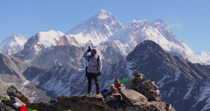 Gokyo Ri Nepal, orbit drone shot of a lone trekker standing among prayer flags on the rocky summit viewpoint looking toward Cholatse peak, surrounded by Everest Region peaks