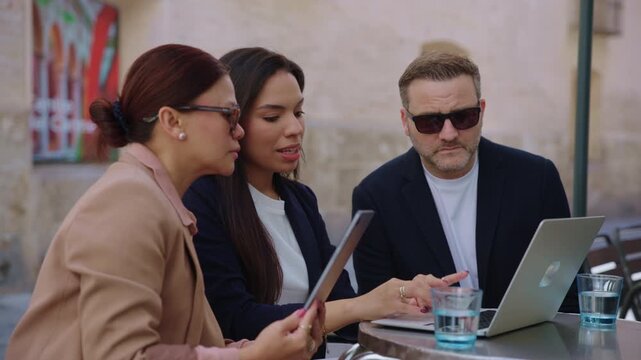 A female leader explains a project on a laptop while a male colleague in sunglasses asks a question during a diverse team meeting at an outdoor cafe.