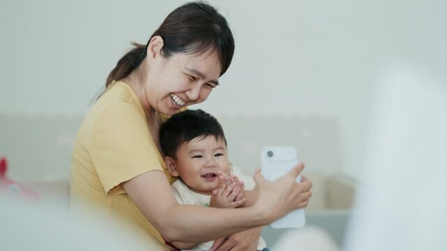 4K Video of a happy Asian mother and her baby boy taking a selfie on a smartphone at home.