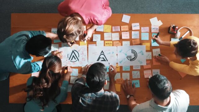 Top down aerial view of skilled designer hold logo while discuss with team. Aerial business group working together sharing idea about logo graphic design by using sticky notes and tablet. Symposium.