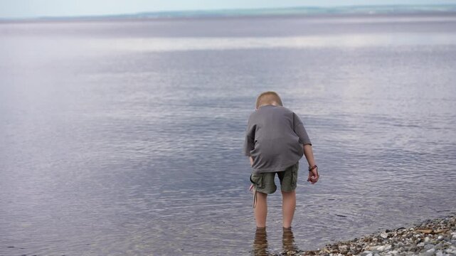A young boy stands at the tranquil water's edge, clad in a grey shirt and green shorts.
