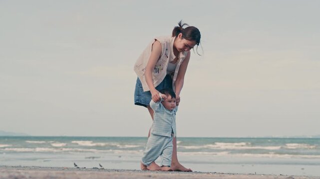 4K Video of an Asian mother holding her baby boy's hands, supporting his first steps on the beach.