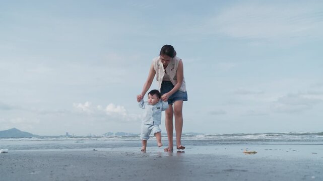 4K Video of an Asian mother holding her baby boy's hands, supporting his first steps on the beach.