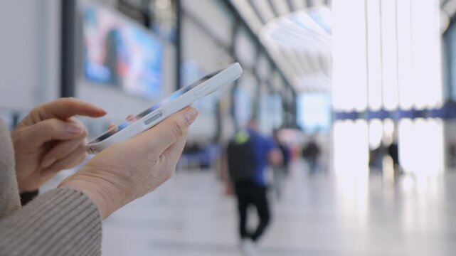 Hand holds smartphone in transit space. Background shows blurred travelers and bright lights. Person appears focused on device, calm and intent. Ideal for travel, technology, or communication themes