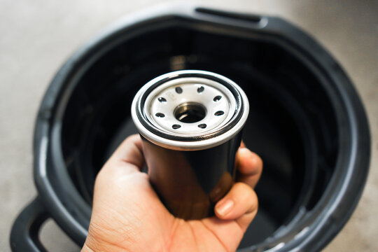 Closeup of hand holding a new black car oil filter with rubber gasket over an oil drain pan during an engine oil change service