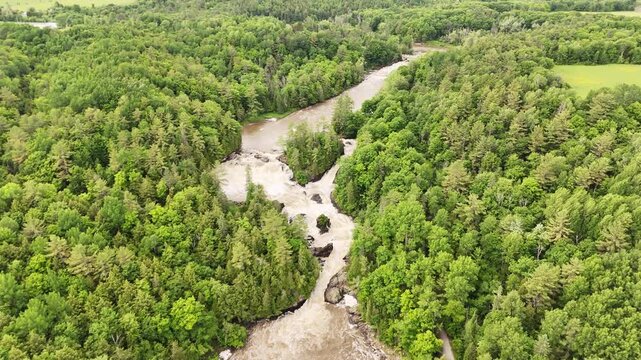 Drone flies forward over lush green forest toward cascading waterfall at Plaisance Park, Quebec. High spring water flow, aerial perspective.