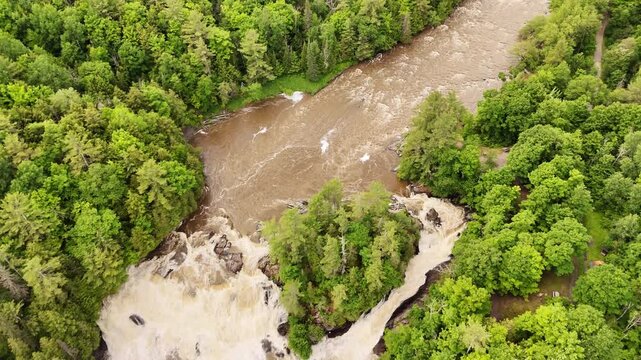 Drone gradually tilts up revealing landscape around Chutes de Plaisance Waterfalls, Quebec Park. Spring high water, cascading falls, lush green forest, aerial overhead perspective.