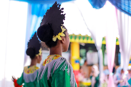 profile and back view of a young female dancer wearing a green traditional costume and a prominent black headdress adorned with a yellow flower ornament.