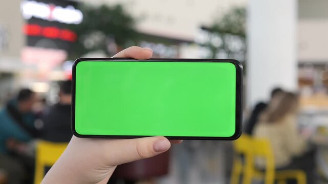 Hand holds phone with green screen. Background shows blurred mall scene. People sit at tables, unaware of camera. Ideal for app ads or tech presentations. Green screen allows easy content replacement