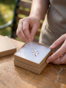 Hands revealing silver leaf pendant from natural kraft gift box on wooden surface, thoughtful present concept