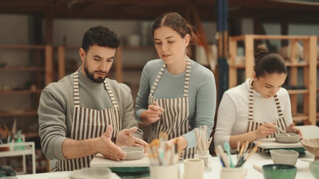 Man craftsperson in striped aprons busy molding clay products in lesson with girl teacher. Amateur sculpting plate of raw clay under supervision of teacher