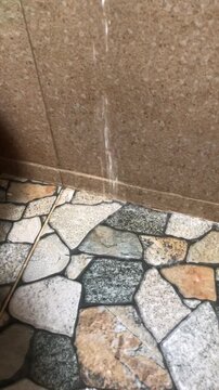 A vertical stream of water trickles continuously down a beige tiled bathroom wall, splashing onto a stone-patterned floor. The motion captures a clear, flowing liquid leak in a modern bathroom.