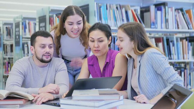 Cheerful young adult university students studying at library using books and laptop, talking and smiling while working on group project