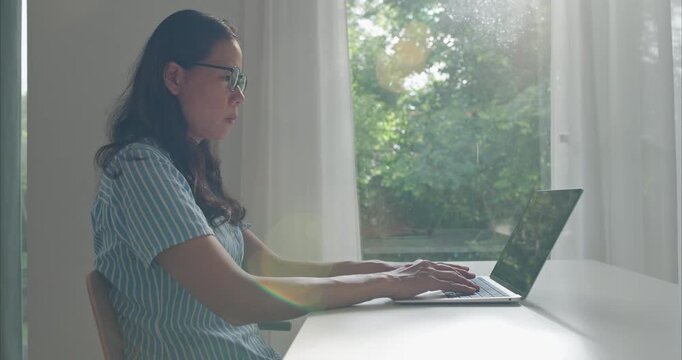 Asian middle female wear glasses typing working on computer, Asia business woman using  laptop at home office morning sunlight, Side view.	