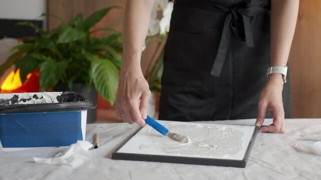 Woman makes relief artwork on canvas shaping white plaster paste with a palette knife on table at home. Concept of DIY textured wall art and creative hobby