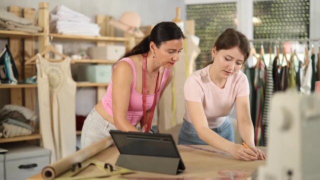 Using ruler and chalk, women tailor draws dress diagram scheme on paper, tablet on table. Craftsman creates pattern for making dress. 