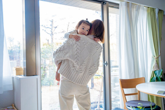 Back view of an Asian mother in a white knit sweater carrying her toddler while looking out a large window