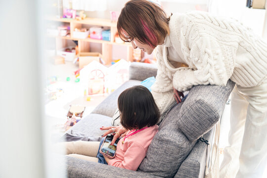 Candid side view of a loving Asian mother watching her little daughter use a smartphone on a sofa
