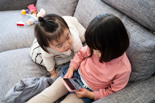 High angle view of two little Asian children sharing a smartphone screen together on a grey sofa
