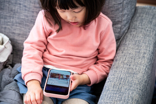 Close up portrait of a concentrated little Asian girl holding a smartphone on her lap