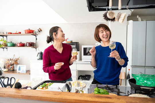 Two cheerful Asian women laughing heartily and holding wine glasses in a modern kitchen